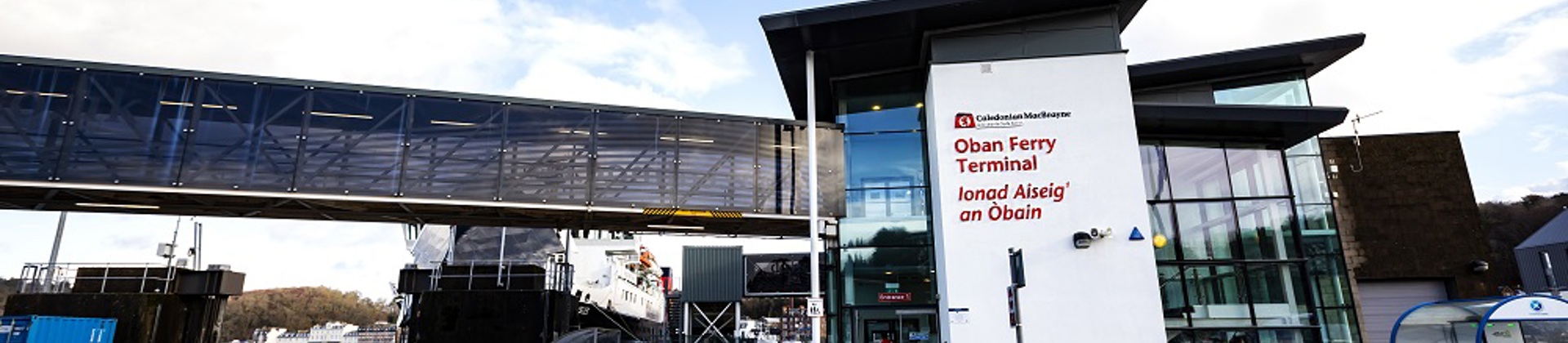 Oban Ferry Terminal entrance with English and Gaelic signage, a skybridge, and a ferry docked under a partly cloudy sky.